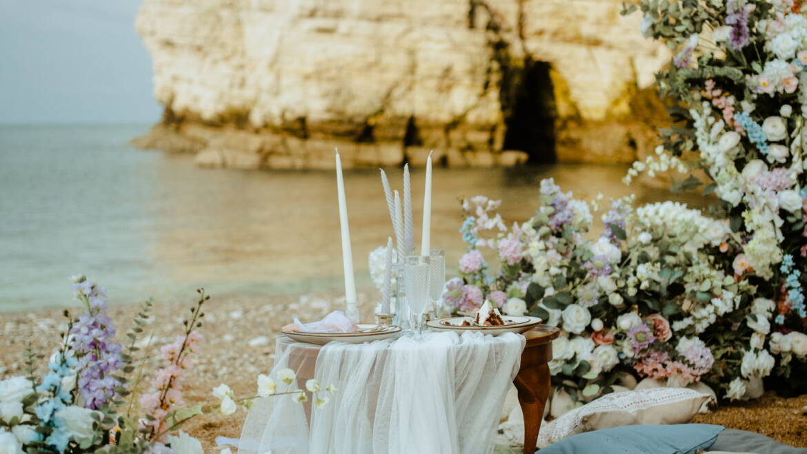 Romantic seaside elopement picnic with soft pastel florals, flowing linens, and candles set against coastal Yorkshire cliffs