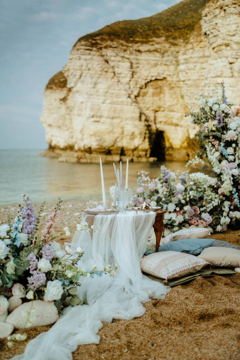 Romantic seaside elopement picnic with soft pastel florals, flowing linens, and candles set against coastal Yorkshire cliffs